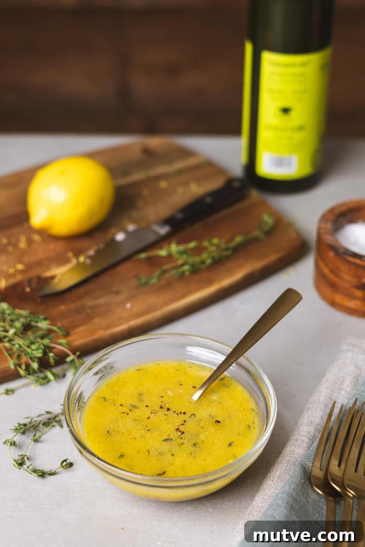 A gray table with a bowl of lemon thyme vinaigrette in the center, fresh thyme leaves and lemon on a cutting board in the background, and a bottle of oil in the background.