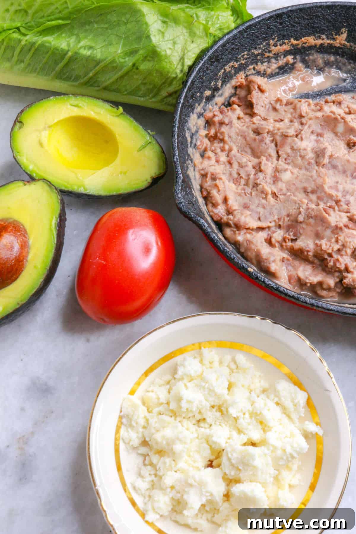 Traditional Mexican Refried Bean Sopes Recipe 3 Lettuce, a cut avocado, a Roma tomato, a skillet with refried beans and a bowl with crumbled queso fresco