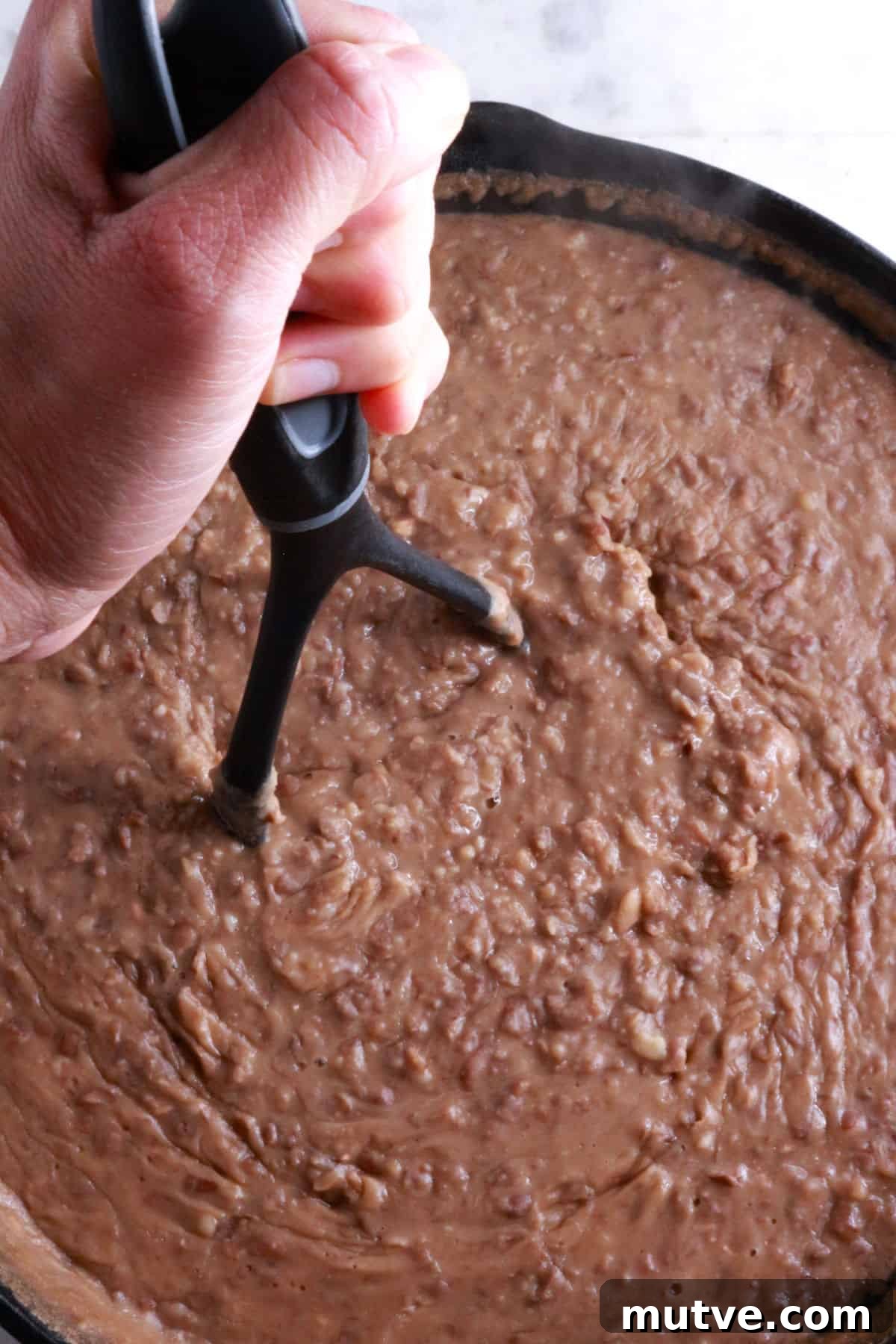 Traditional Mexican Refried Bean Sopes Recipe 4 A hand on a potato masher mashing beans in a cast-iron skillet