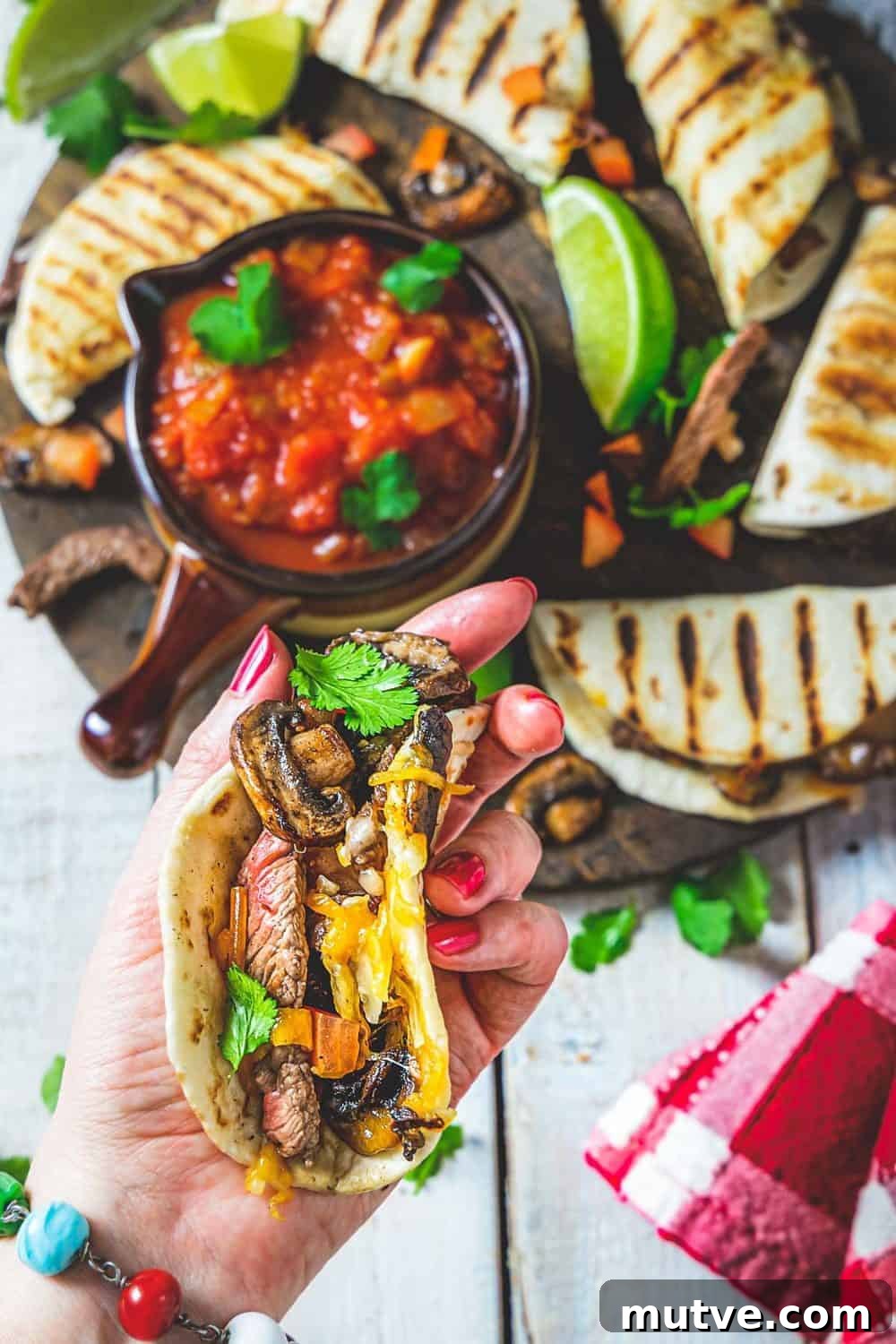 A person holding a mini tortilla filled with steak strips, mushrooms and cheese.