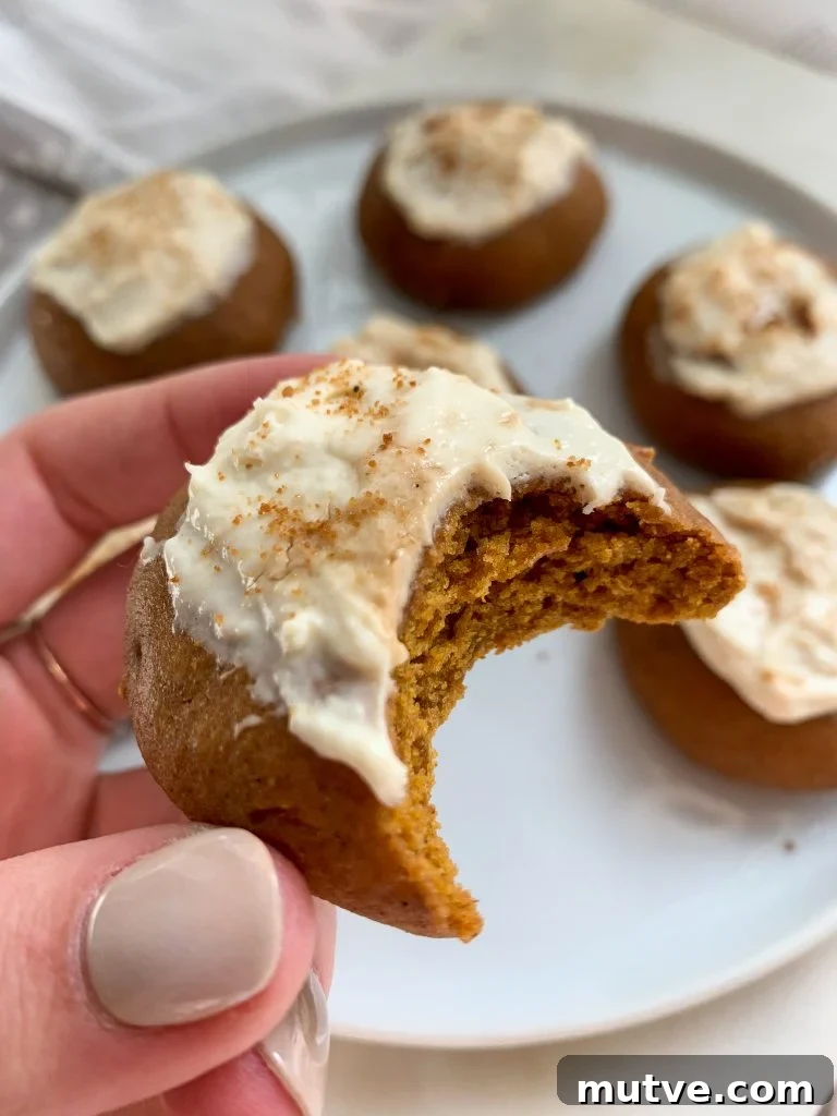 a hand holding a whole wheat pumpkin cookie with a maple cream cheese frosting over a white plate