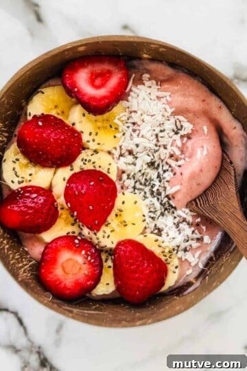 A spoon in the strawberry banana smoothie bowl that is topped with strawberries, banana, coconut, and chia seeds.