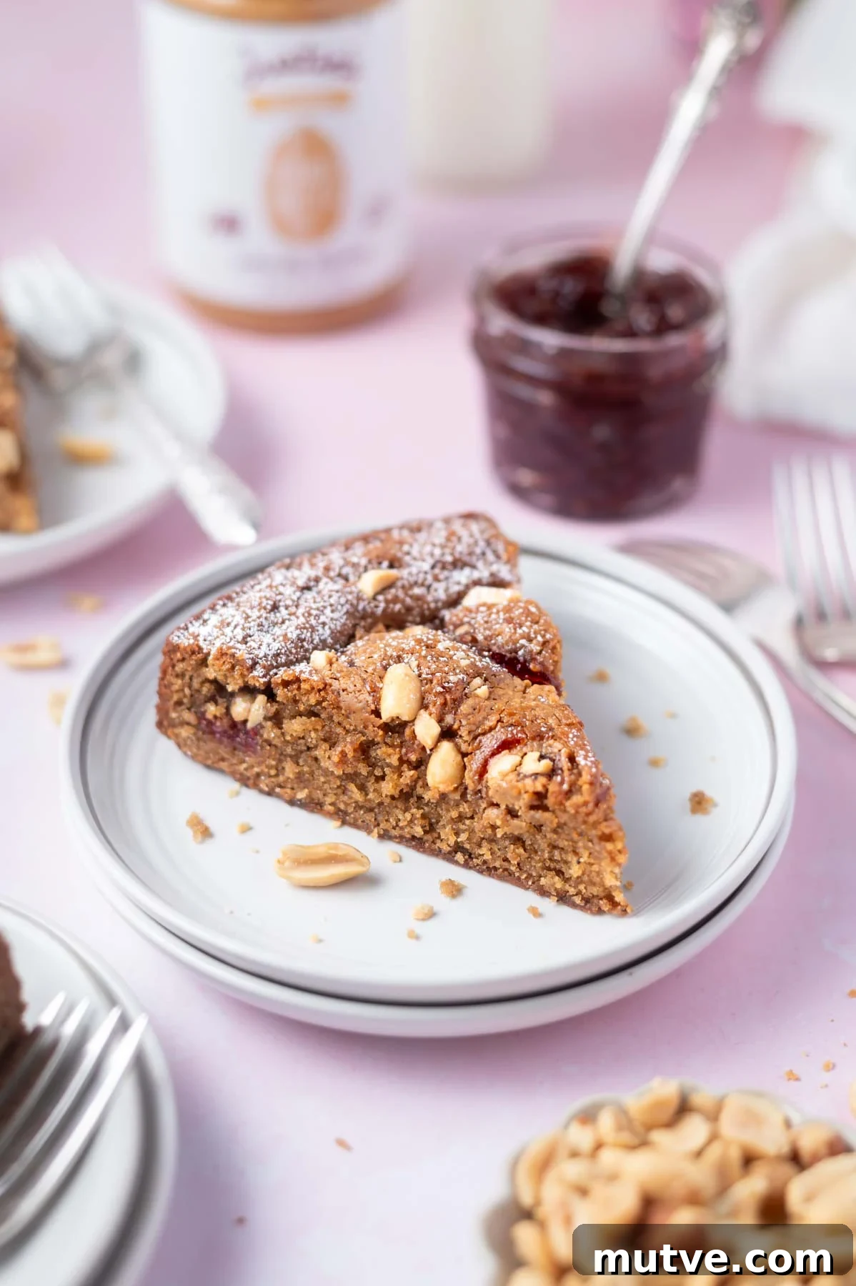 slice of cake on a plate with a jar of jam in the back ground and a bowl of peanuts