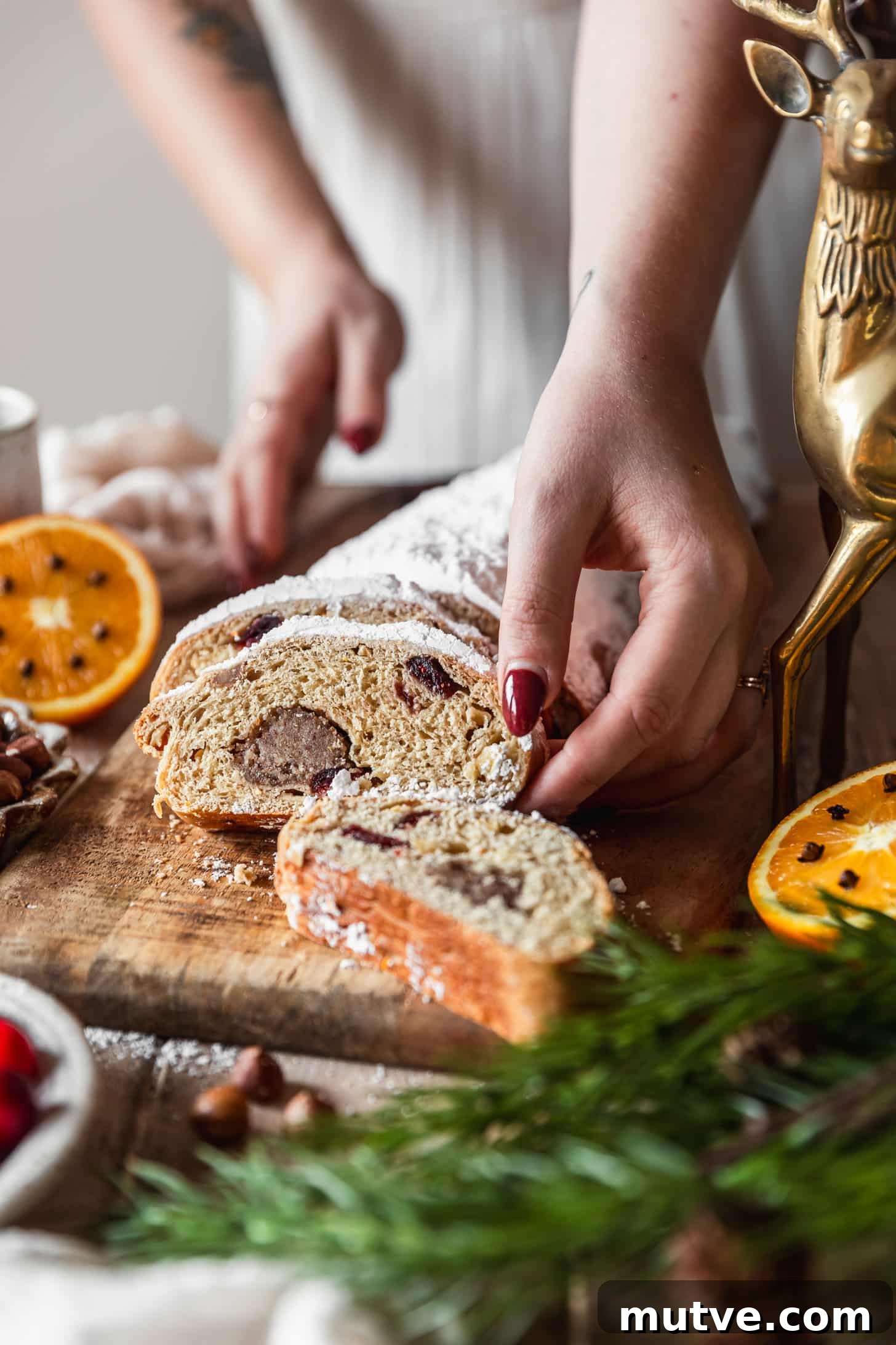 A woman in a white dress reaching for a piece of German Christmas bread on a wood board next to oranges, a gold deer, garland, and hazelnuts.