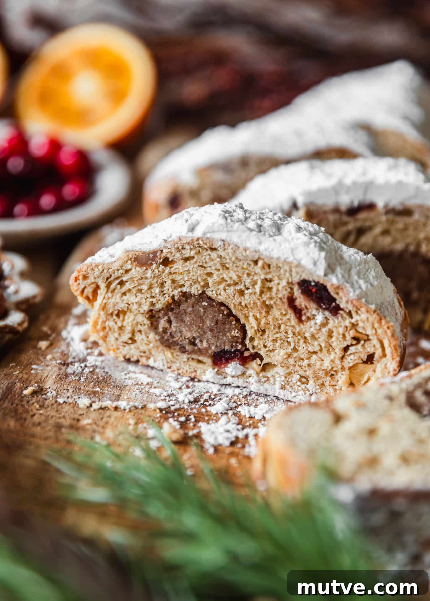 Slices of cranberry apple stollen on a wood board next to oranges, a white bowl of cranberries, and garland.