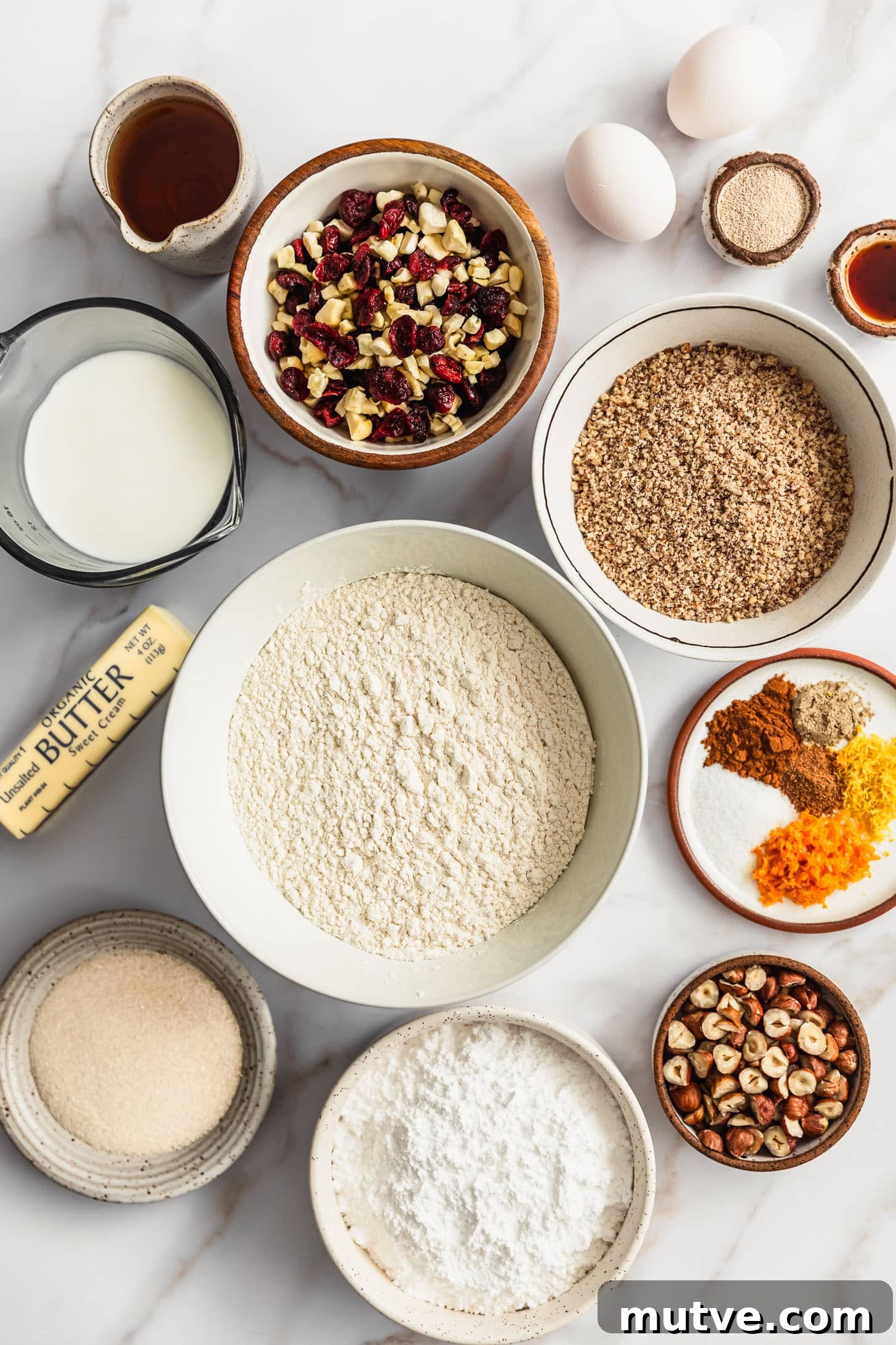 White and brown bowls of flour, hazelnut flour, dried fruit, bourbon, yeast, vanilla, milk, spices, citrus zest, sugar, powdered sugar, and hazelnuts on a white counter next to butter and eggs.
