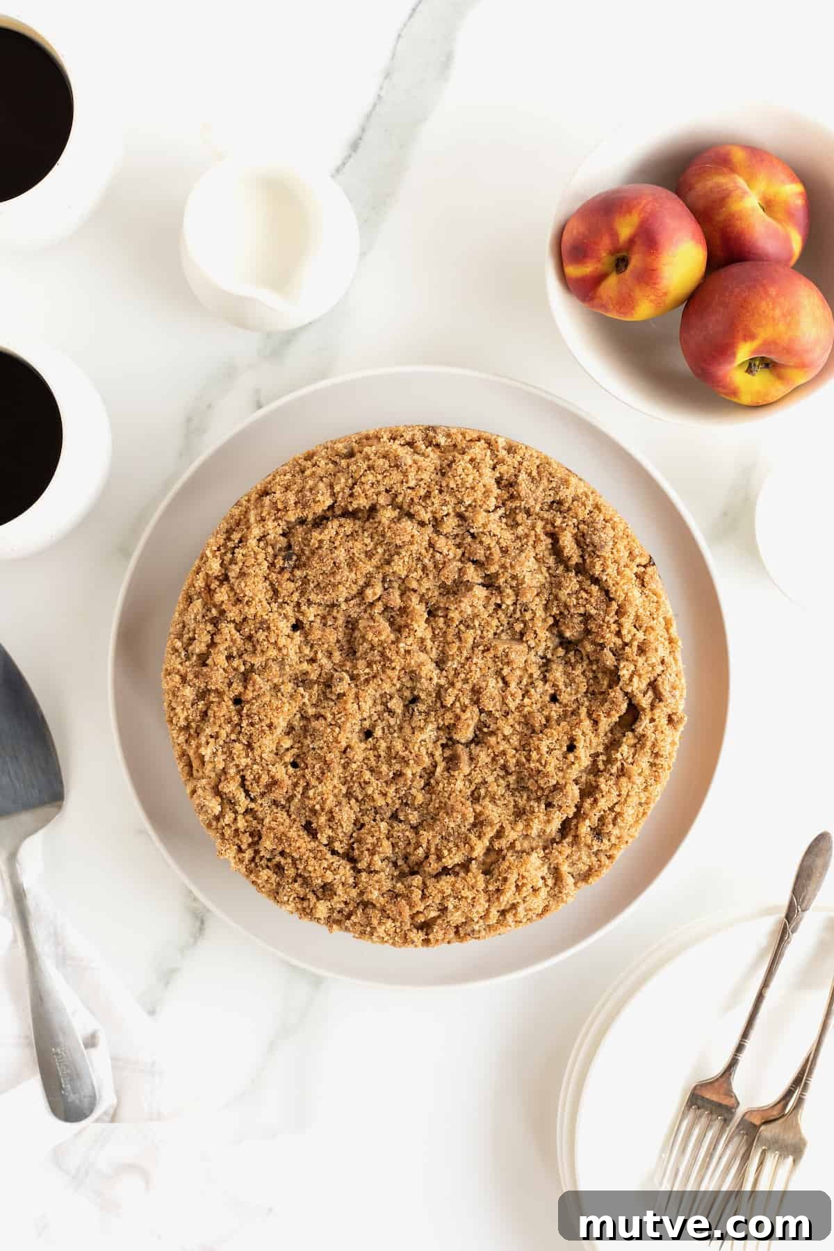 Peach Coffee Cake with Cinnamon Streusel 9 A peach coffee cake with cinnamon sugar streusel on a white serving platter next to a bowl of ripe peaches.