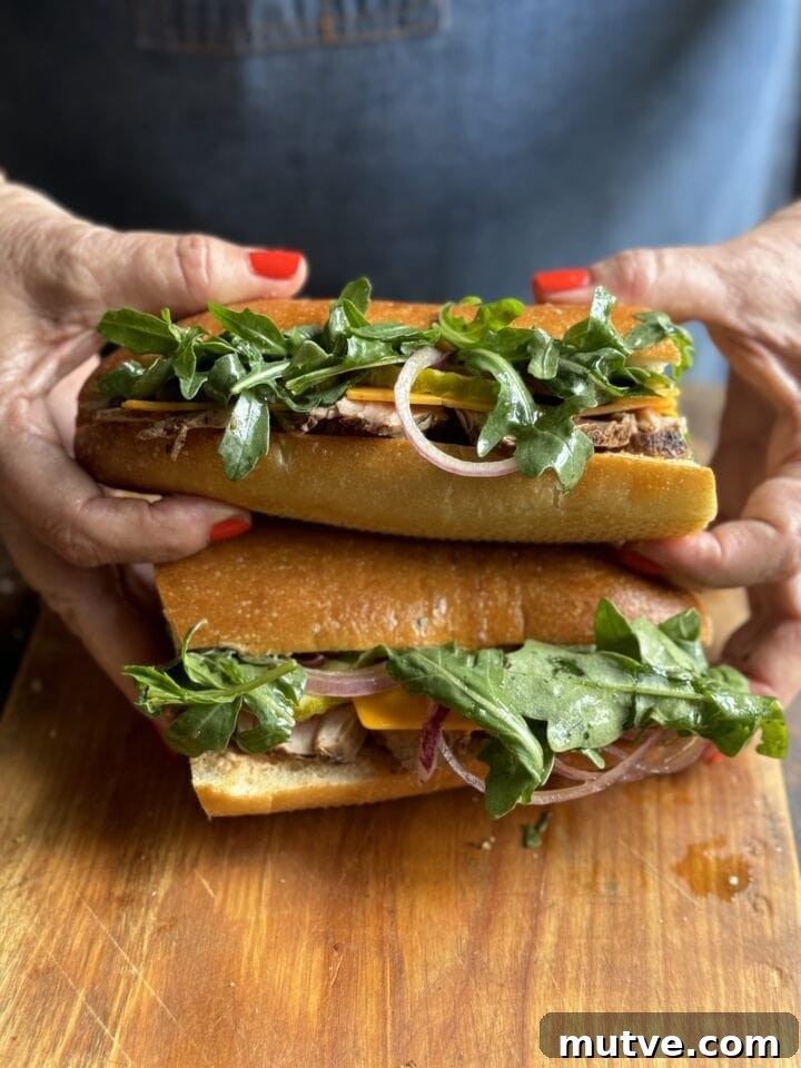 two pork tenderloin sandwiches with arugula cheese and onions on a French roll halves stacked by two hands with red nail polish