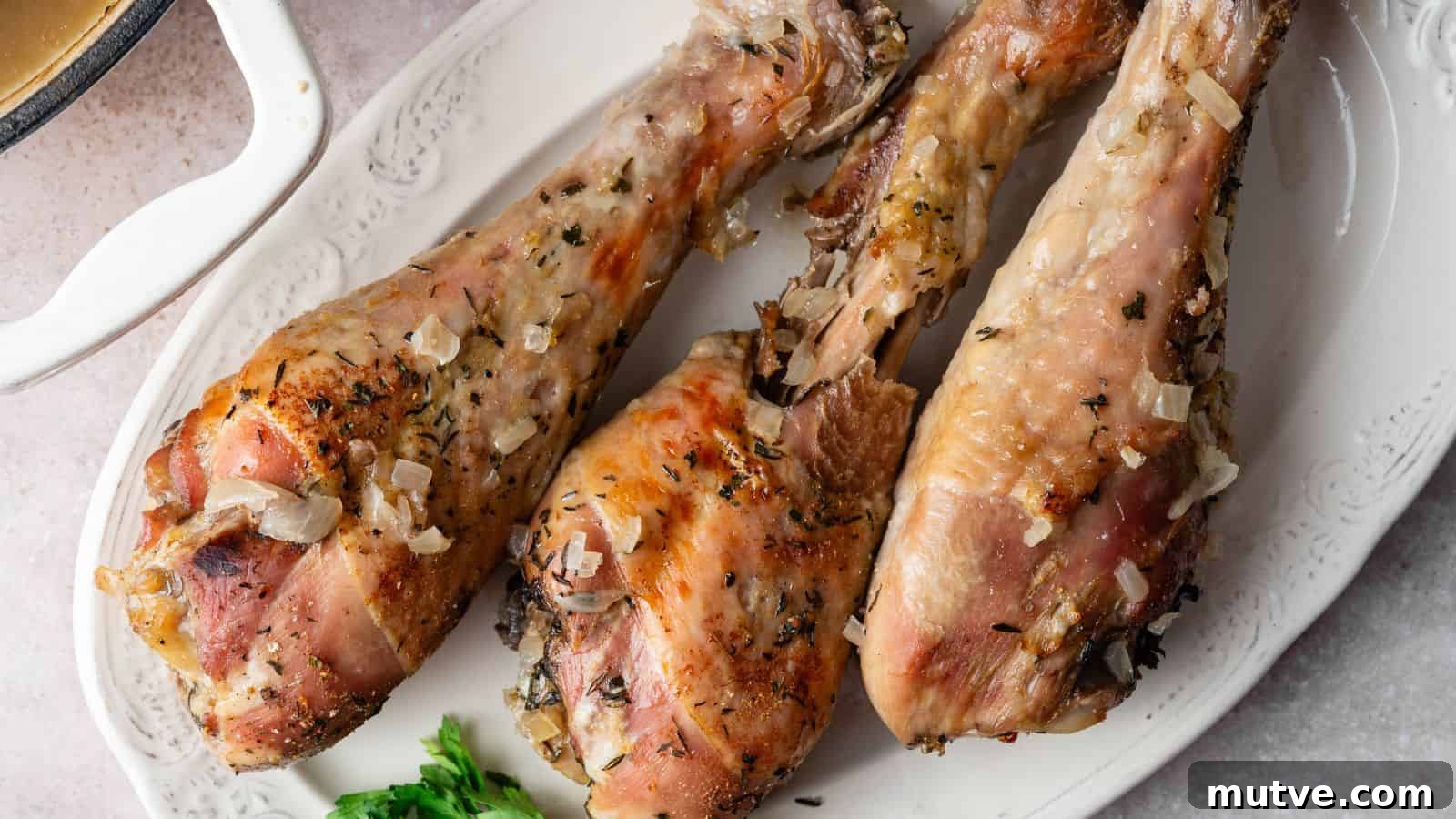 Three cooked turkey legs with herbs and chopped onions are arranged on a white oval platter, garnished with a sprig of parsley.
