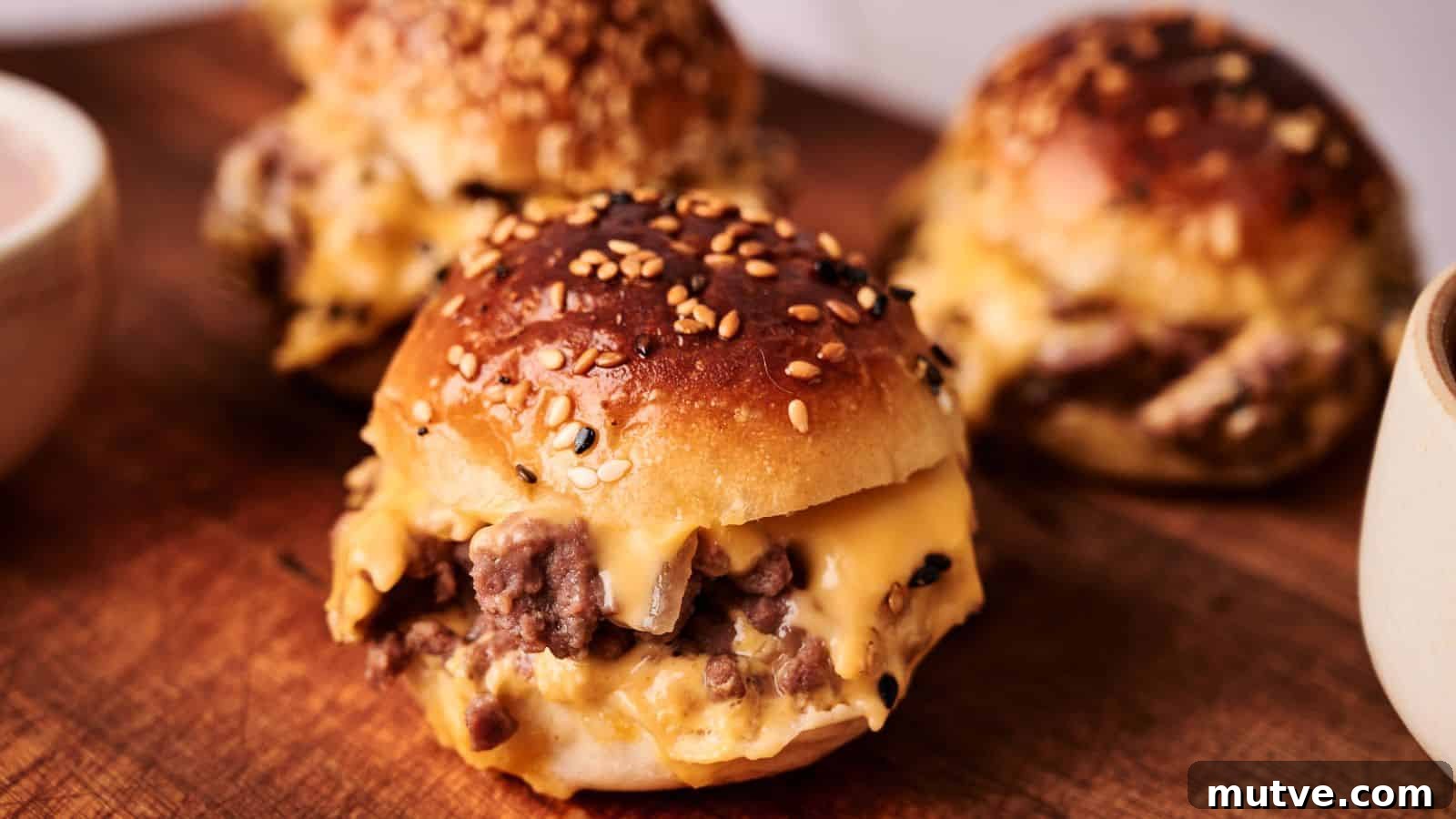 Three sesame seed buns filled with melted cheese and ground beef are arranged on a wooden board.