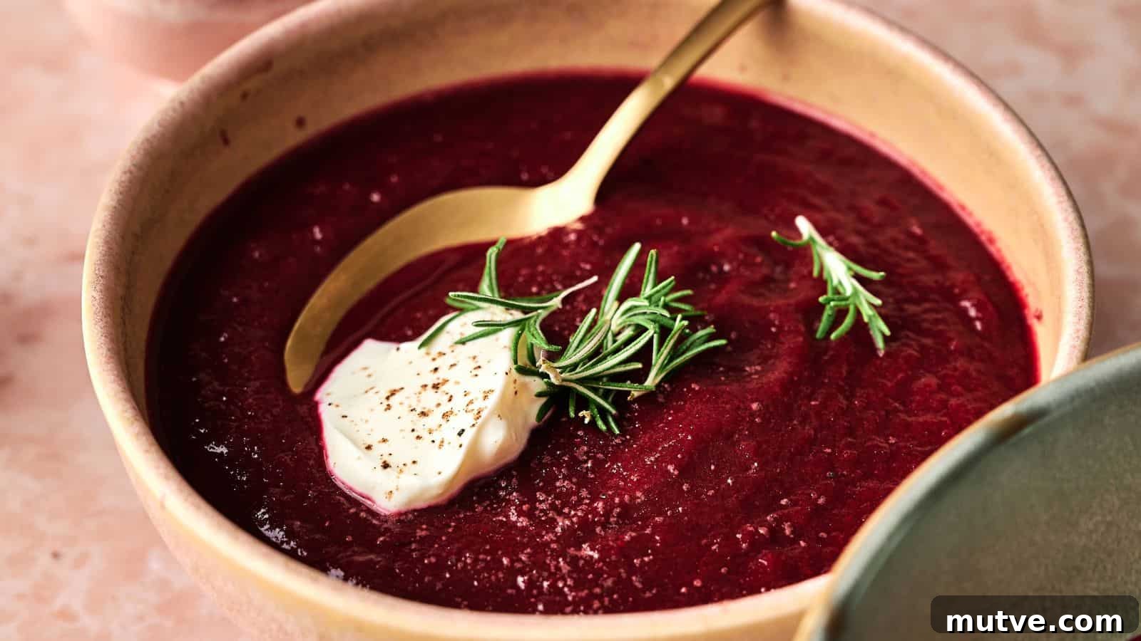 A bowl of beet soup is garnished with a dollop of cream, black pepper, and fresh rosemary, with a gold spoon resting inside the bowl.
