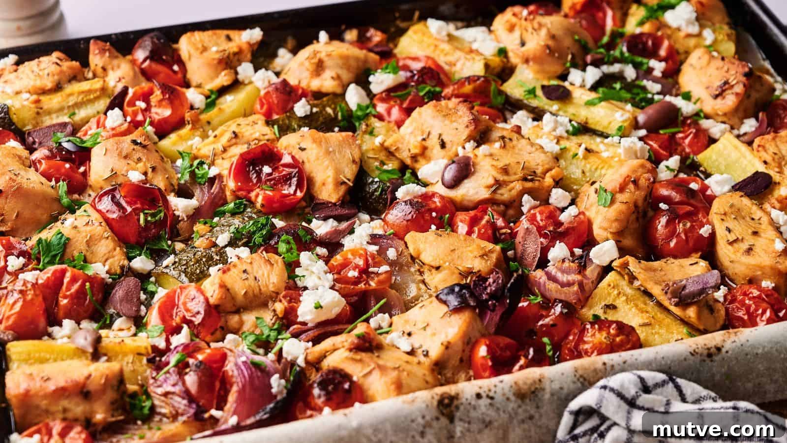 Sheet pan filled with baked chicken pieces, cherry tomatoes, zucchini, olives, and crumbled feta cheese, garnished with herbs.