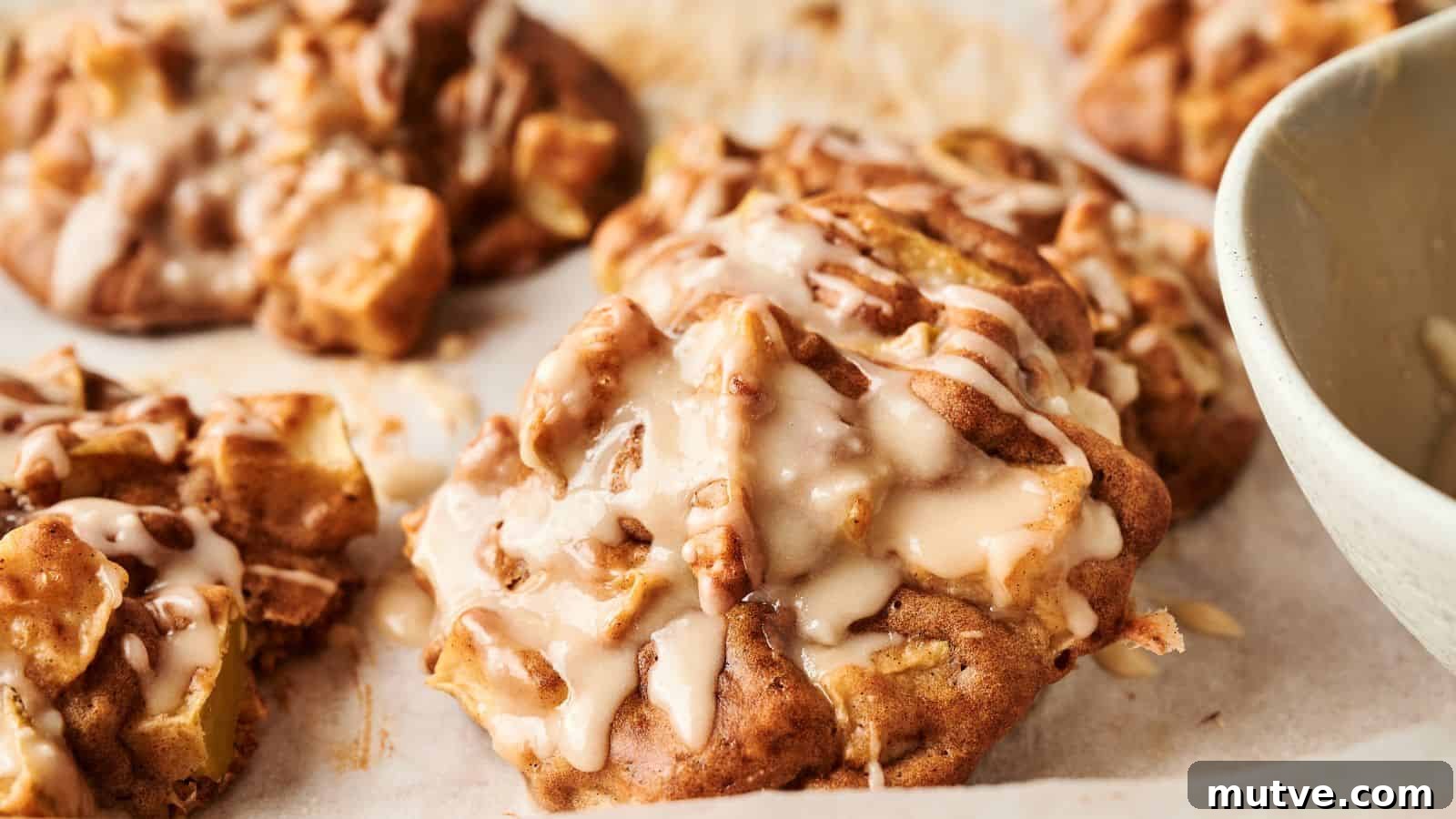 Close-up of freshly baked apple cookies drizzled with icing, resting on parchment paper with part of a bowl visible to the right.