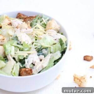 One chicken Caesar salad in a white bowl with croutons to the right of the bowl.