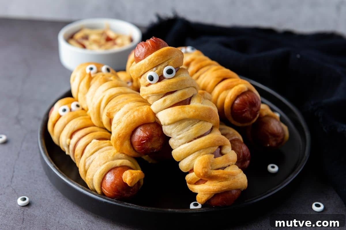 29 Halloween Treats That Beat Your Trick-or-Treat Candy 12 Hot dogs wrapped in crescent dough to resemble mummies, topped with candy eyes, arranged on a black plate with a small bowl of dipping sauce in the background.