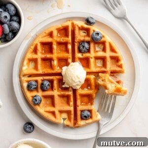 Overhead view of a gluten-free Belgian waffle on a white plate topped with whipped butter, maple syrup, and fresh blueberries, with a bite taken out on a fork.