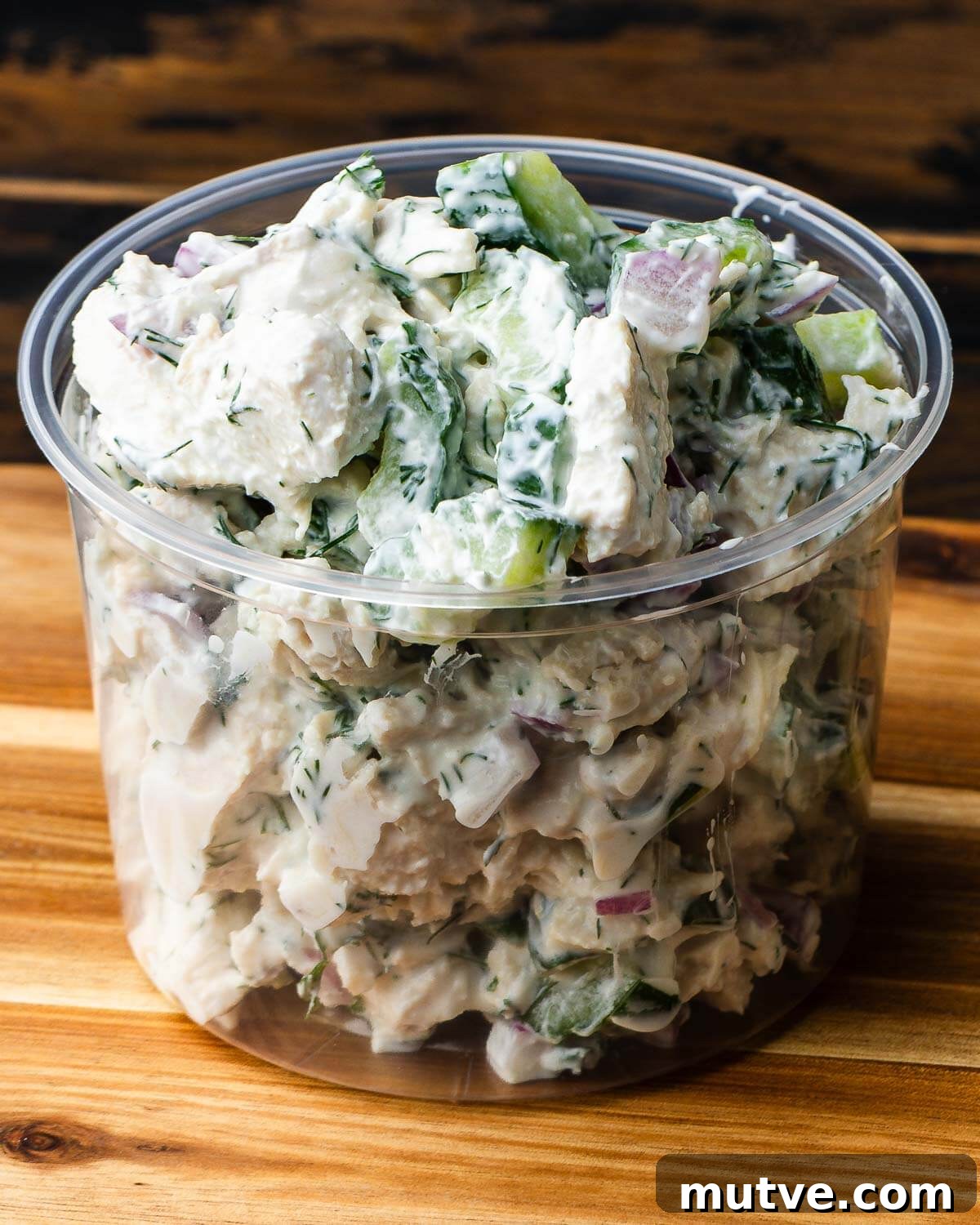 Creamy chicken cucumber salad in deli container on wooden board.