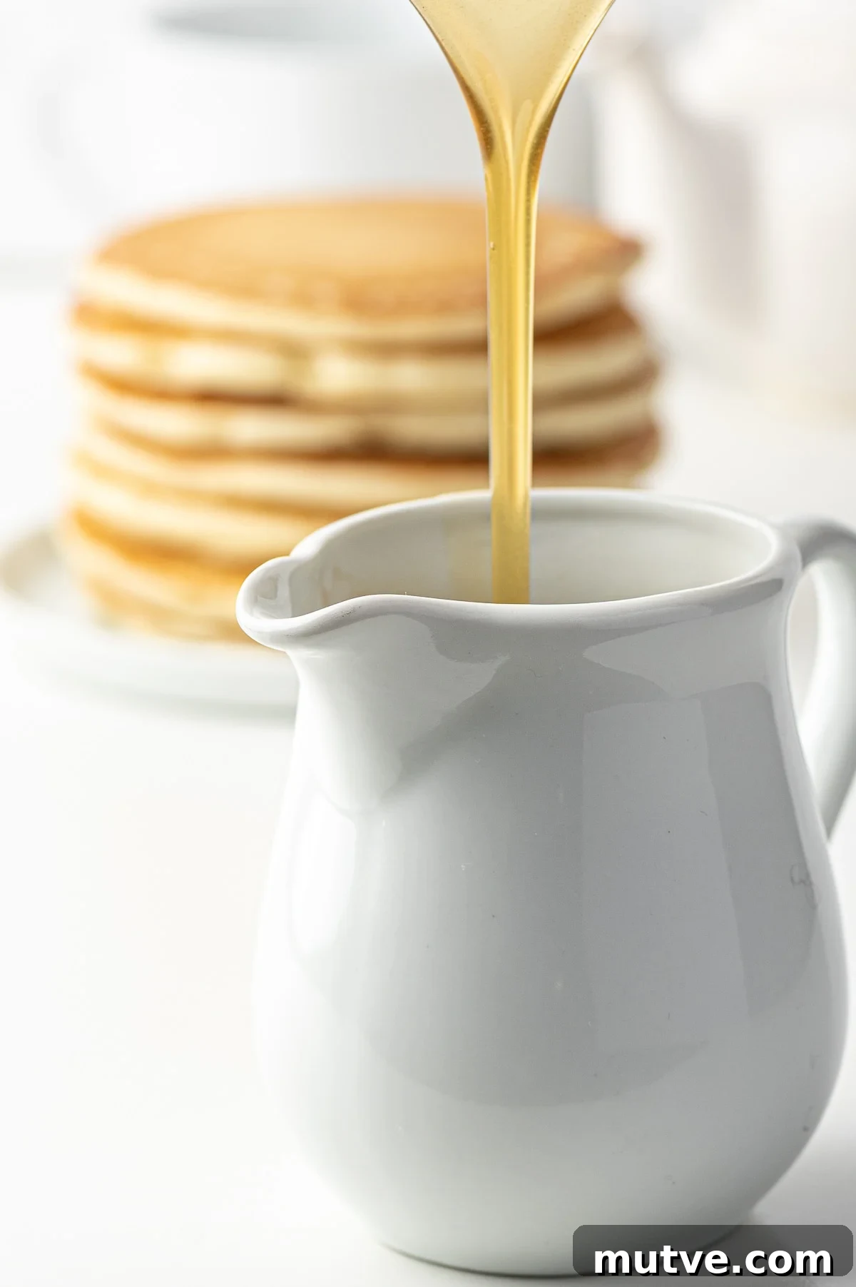 How to Make Keto Maple Syrup That Won't Crystallize 3 A stream of golden keto pancake syrup being poured into a white dish. A stack of pancakes in the background slightly out of focus.