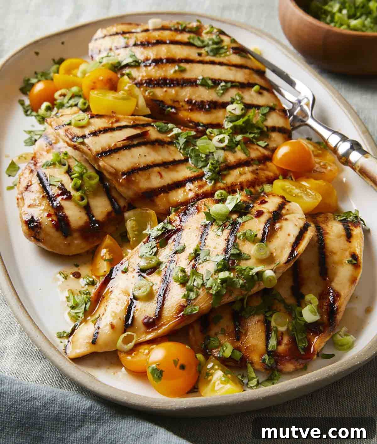 Five chicken breast pieces with grill marks on a platter, topped with halved cherry tomatoes, scallions, and cilantro.