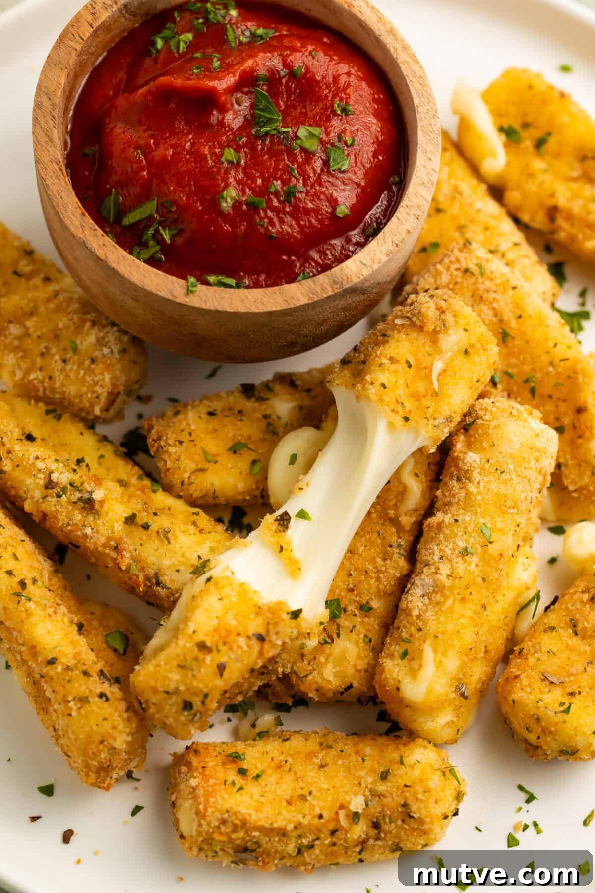 Air Fryer Mozzarella Sticks with Gluten Free Option 2 Close up of a pile of air fry mozzarella sticks with a wooden bowl of tomato sauce. One of the mozzarella sticks is broken in half to show the cheese pull.