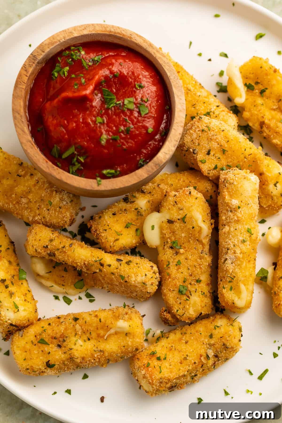 Air Fryer Mozzarella Sticks with Gluten Free Option 3 Close up of a white plate of air fryer mozzarella sticks with a scattering of finely chopped herbs and a wooden bowl of tomato dipping sauce.