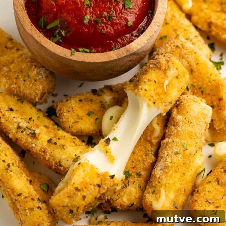 Close up of a pile of air fry mozzarella sticks with a wooden bowl of tomato sauce. One of the mozzarella sticks is broken in half to show the cheese pull.