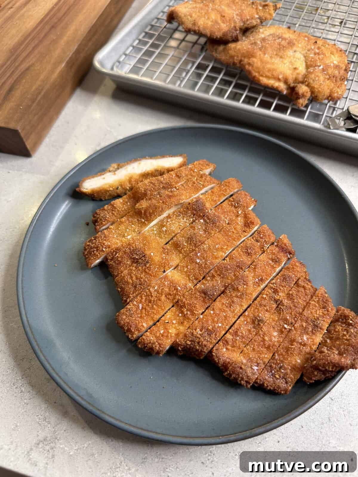 A sliced, golden-brown breaded cutlet is arranged on a dark plate, with additional cutlets cooling on a wire rack in the background.
