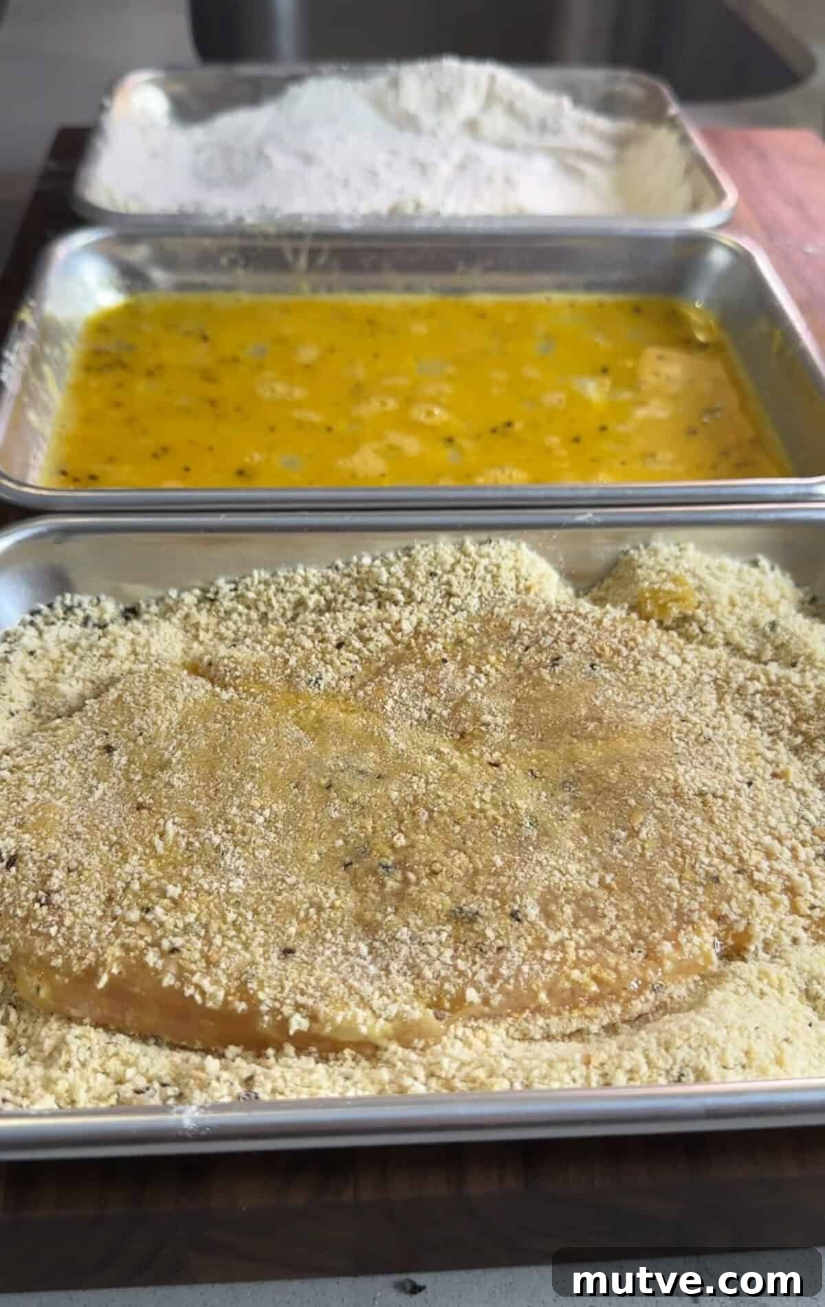 A breaded cutlet is being coated in seasoned breadcrumbs on a tray, with trays of flour and egg wash visible in the background, ready for the breading process.