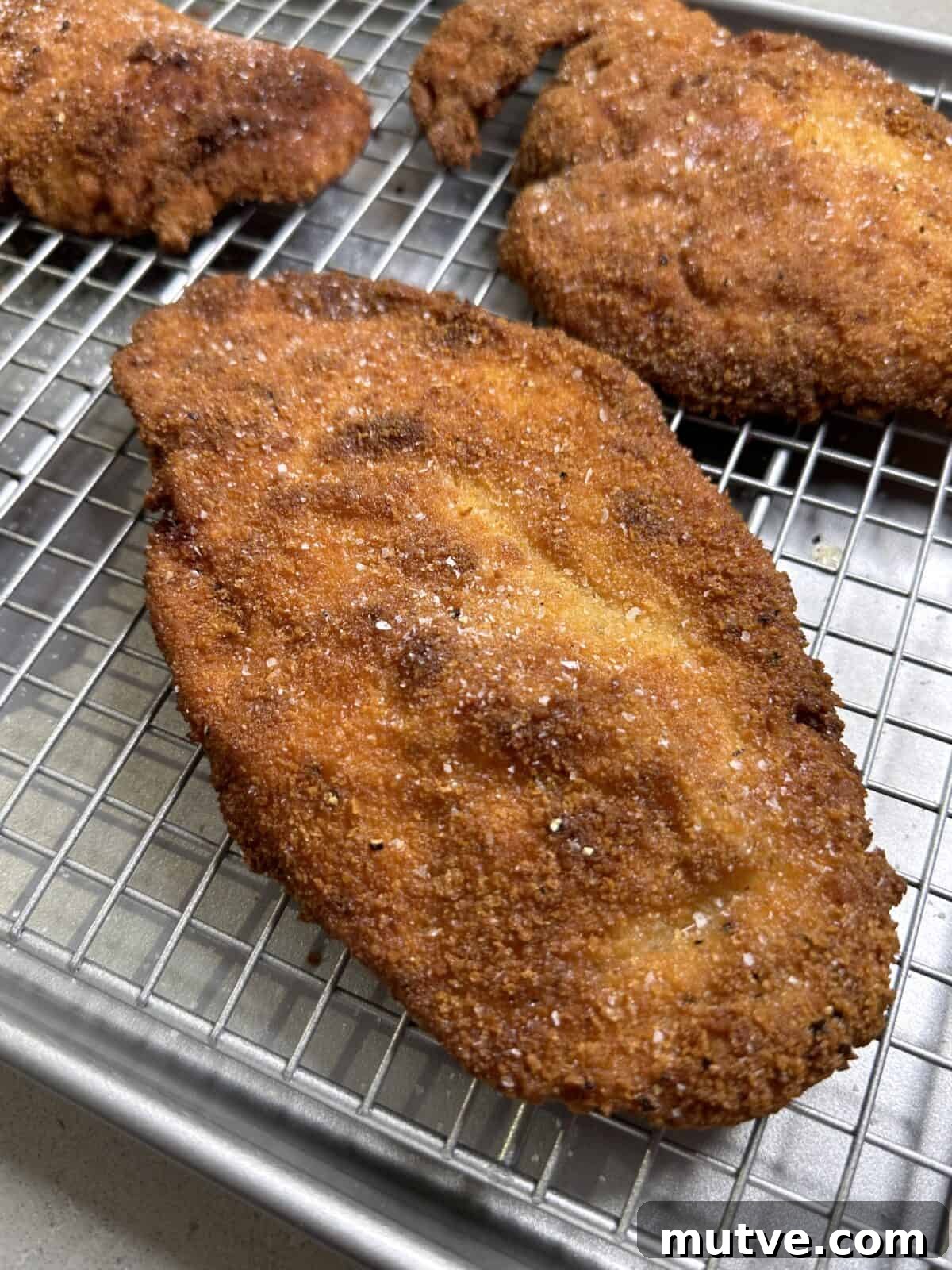 A close-up of a golden-brown breaded and fried chicken cutlet resting on a wire rack over a baking sheet, with a light sprinkle of salt on top.