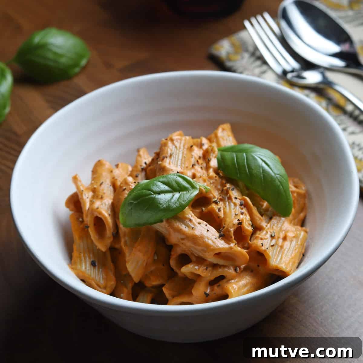 Photo of a bowl of vegan creamy tomato pasta on a wooden tabletop with cutlery and napkin.