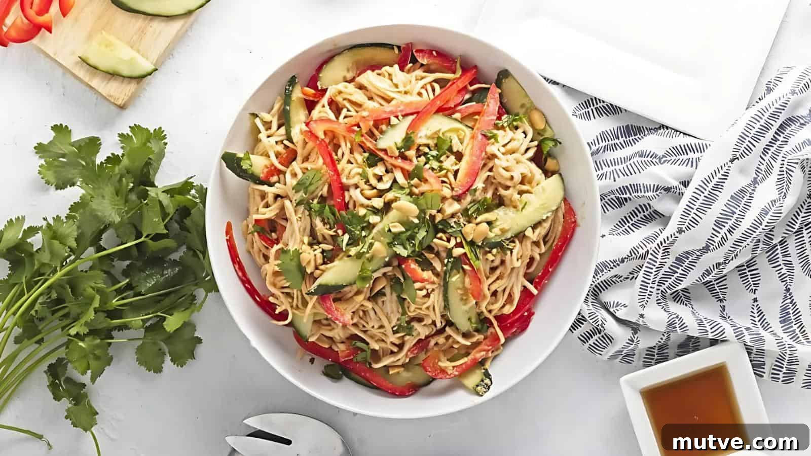 40 End-of-Summer Cookout Recipes to Celebrate Summer's Finale 27 A bowl of noodle salad with sliced cucumbers, red bell peppers, and chopped peanuts, garnished with cilantro, on a white surface with a striped napkin and a small bowl of sauce nearby.