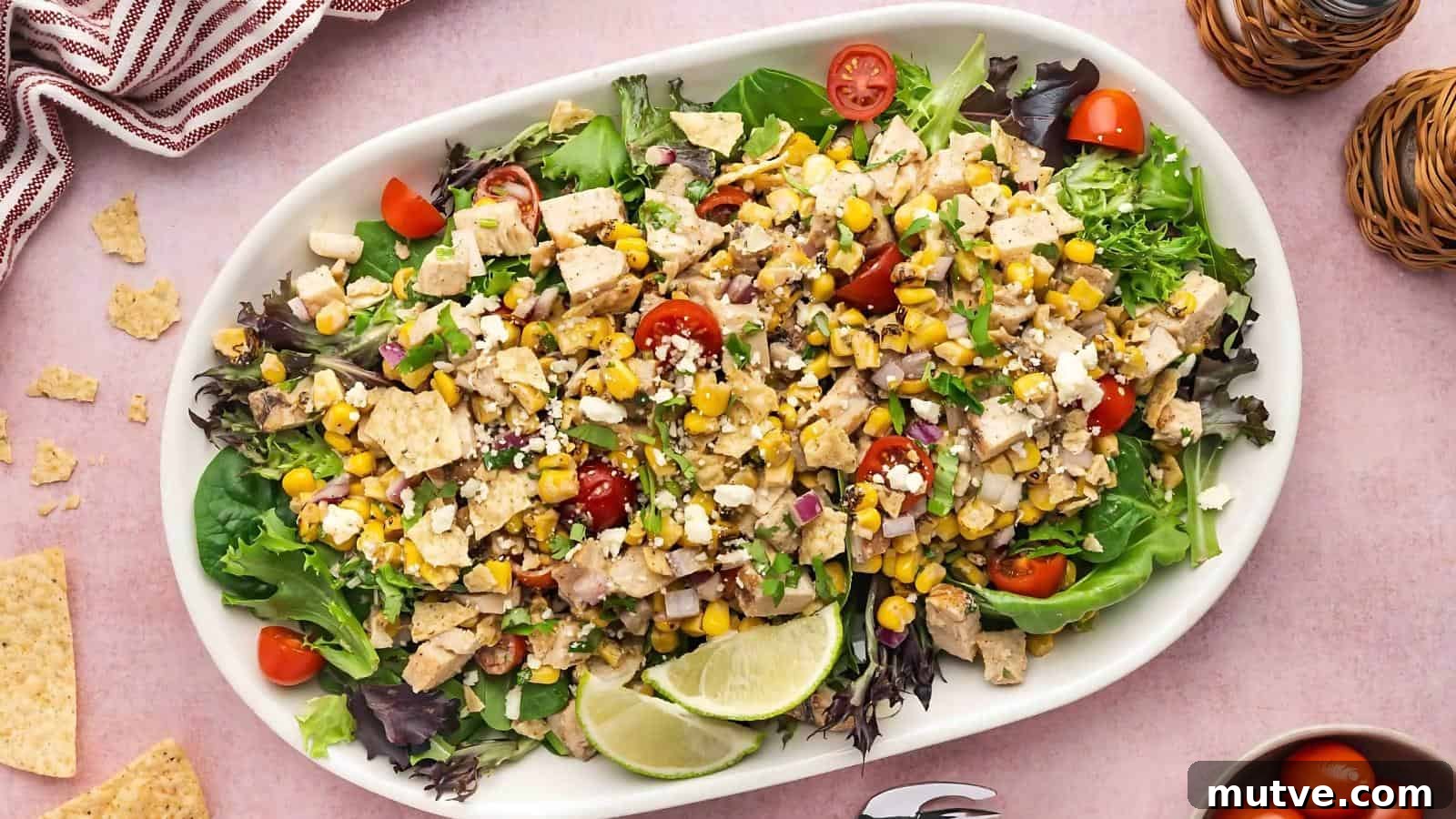 40 End-of-Summer Cookout Recipes to Celebrate Summer's Finale 32 A large white platter filled with chopped salad greens, diced chicken, corn, cherry tomatoes, crumbled cheese, tortilla chips, and lime wedges on a pink surface.