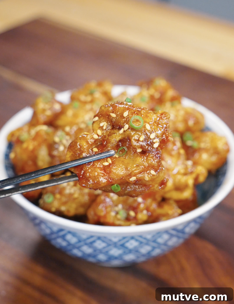 Chopsticks holding a crispy piece of honey butter fried chicken coated in a glossy sweet sauce with sesame seeds and scallions.
