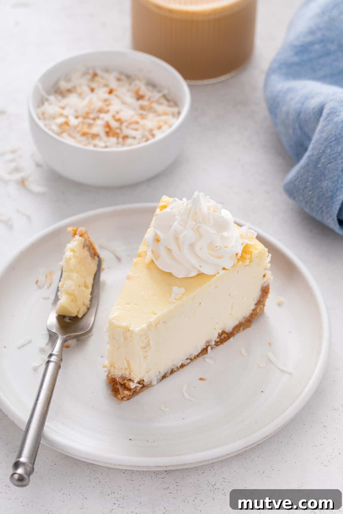 Creamy Coconut Cheesecake Recipe 4 Bite taken from a slice of coconut cheesecake on a white plate.