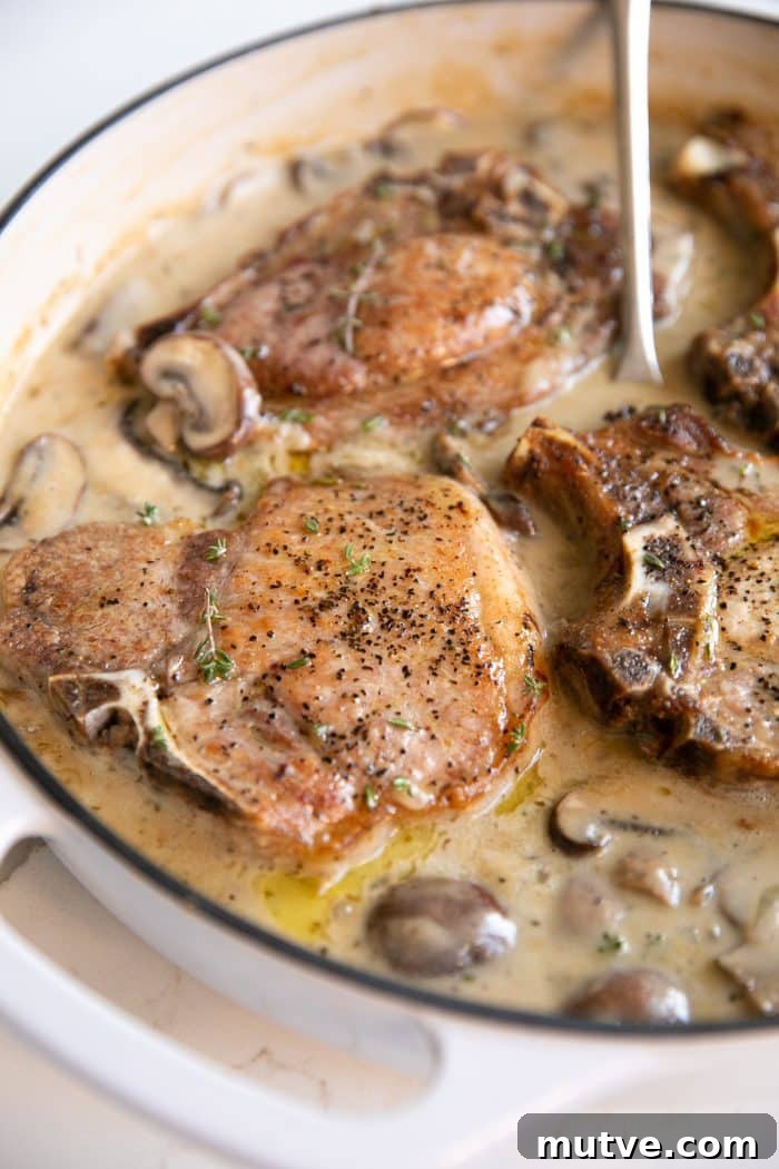 One-Pan Smothered Pork Chops with Mushroom Gravy 5 White ceramic pan filled with cooked bone-in pork chops simmering in creamy mushroom sauce.