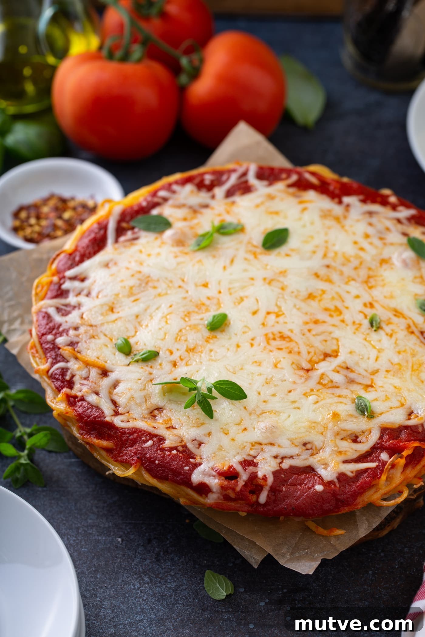 A baked spaghetti pie in a pie dish. There are layers of spaghetti, sauce, and melted cheese. Fresh basil is sprinkled on top and three tomatoes, olive oil, a container of dried red pepper flakes, and dinner plates are next to the spaghetti pie.