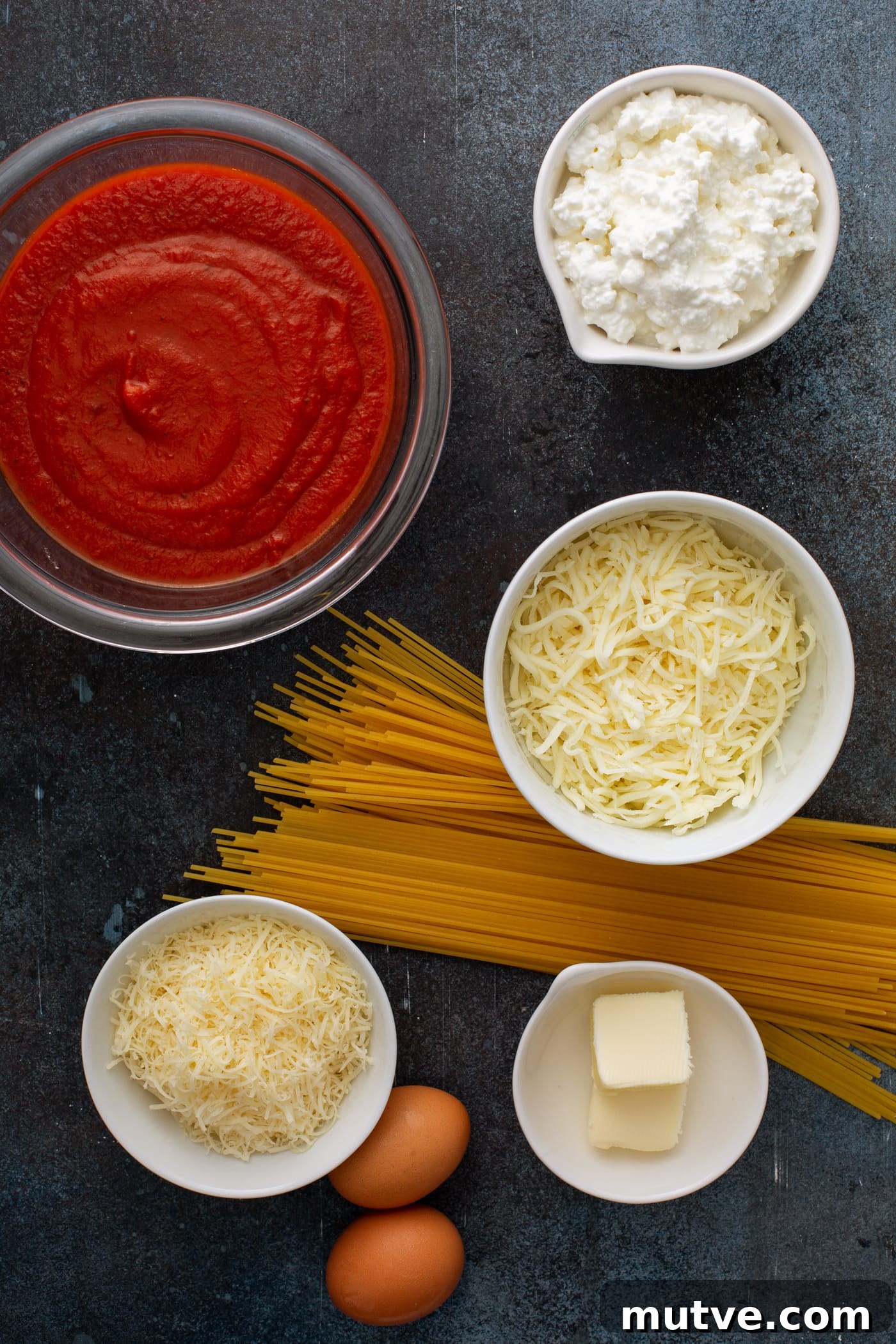 Small bowl containing ingredients for baked spaghetti pie. There is a handful of uncooked spaghetti pasta, a bowl of spaghetti sauce, small containers of shredded mozzarella cheese, cottage cheese, and Parmesan cheese.