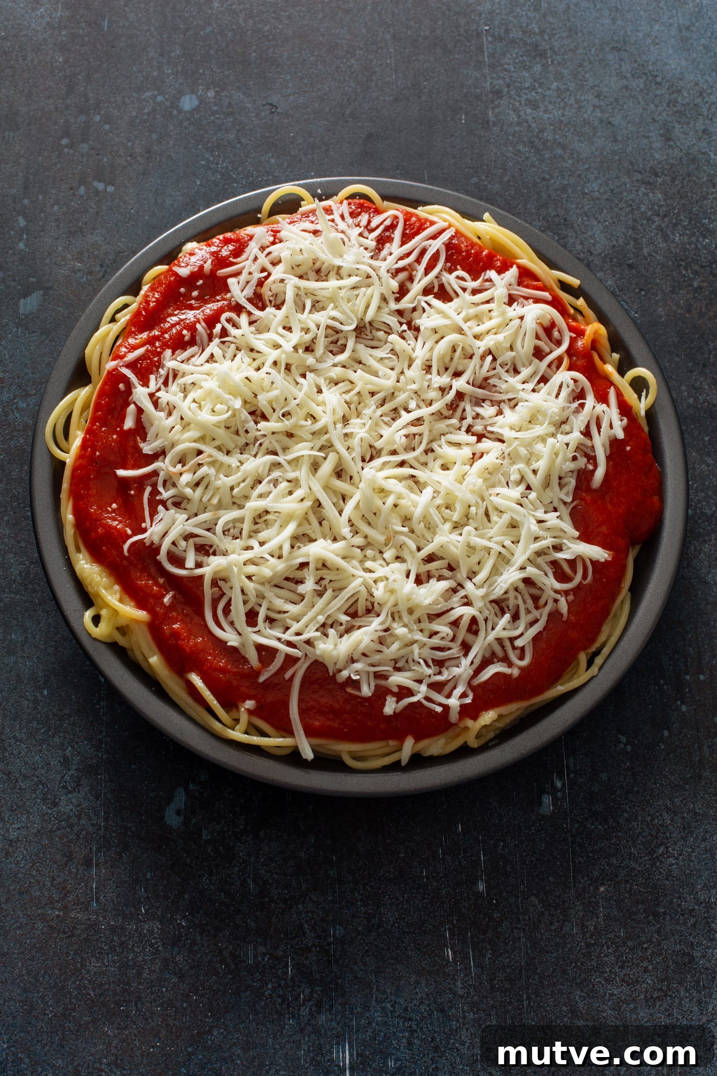 A pie dish filled with cooked spaghetti, topped with spaghetti sauce and shredded cheeses.