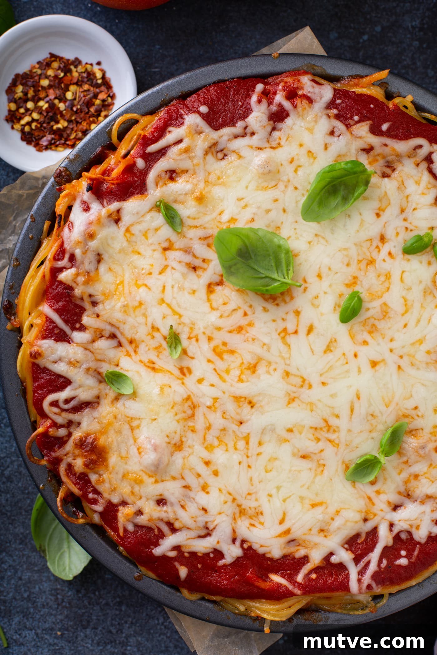 A top view of baked spaghetti pie. There is spaghetti pasta, spaghetti sauce and melted cheeses with a few basil leaves on top. A small bowl of dried pepper flakes is behind the pie.