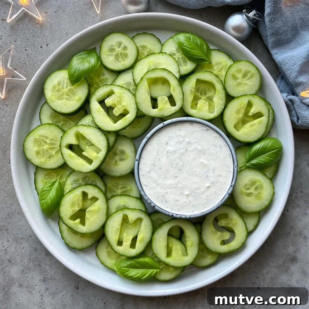 Christmas Veggie Tray Ideas for Holiday Parties 2 A festive Christmas Veggie Tray features cucumber slices with some spelling MERRY XMAS. Fresh basil leaves and a bowl of white dipping sauce sit at the center, surrounded by decorative lights and ornaments.
