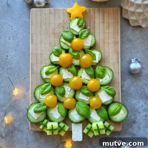 Festive and healthy, this Christmas tree is crafted from cucumber slices, basil, yellow cherry tomatoes, and cheese bites on a wooden board, with little cheese and cucumber presents and a yellow star on top—perfect for holiday food recipes!.