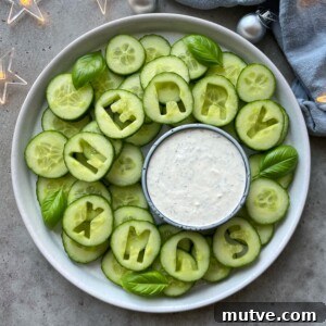 A festive Christmas Veggie Tray features cucumber slices arranged in a wreath, with some spelling MERRY XMAS. Fresh basil leaves and a bowl of white dipping sauce sit at the center, surrounded by decorative lights and ornaments.