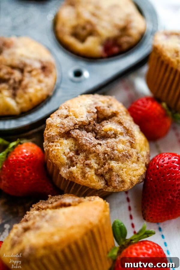 A close up shot of a strawberry rhubarb muffin next to a muffin tin and some fresh strawberries.