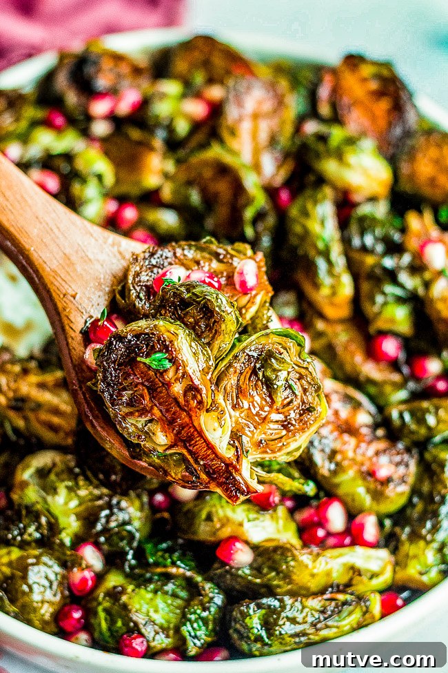 Balsamic Glazed Brussels Sprouts with Pomegranate Seeds 6 Roasted brussels sprouts garnished with pomegranate seeds in a serving bowl with a wooden spoon.