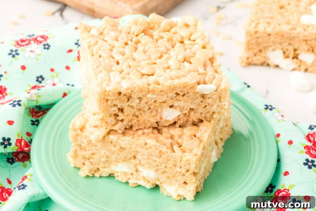 Browned Butter Rice Krispie Treats 13 Close-up of two Browned Butter Rice Crispy Treats stacked on a green plate with a floral cloth and extra bars in the background.