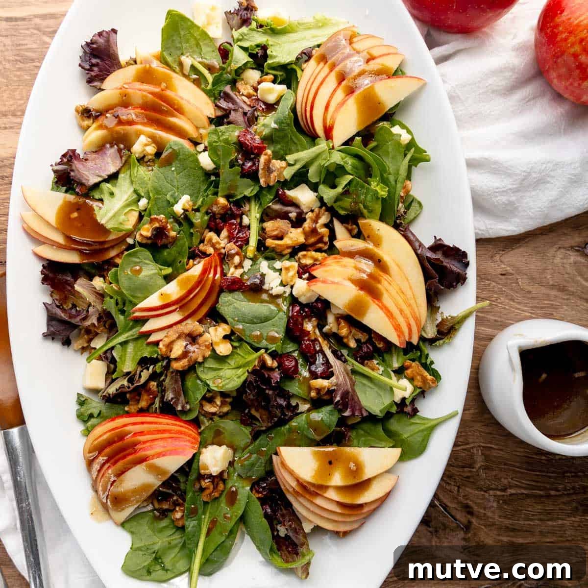 A white oval plate with a mixed greens salad topped with apple slices, walnuts, cranberries, and bits of cheese. A small white pitcher with dressing is on the side. Two red apples and a napkin are in the background.