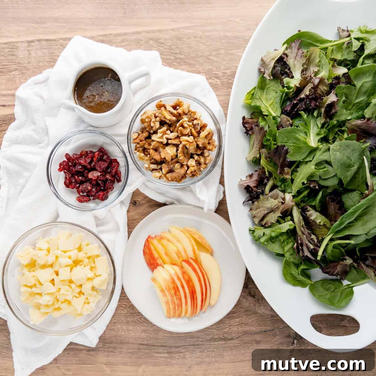 A large bowl of mixed greens with bowls of dressing, walnuts, dried cranberries, cheese, and apple slices arranged on a wooden surface.