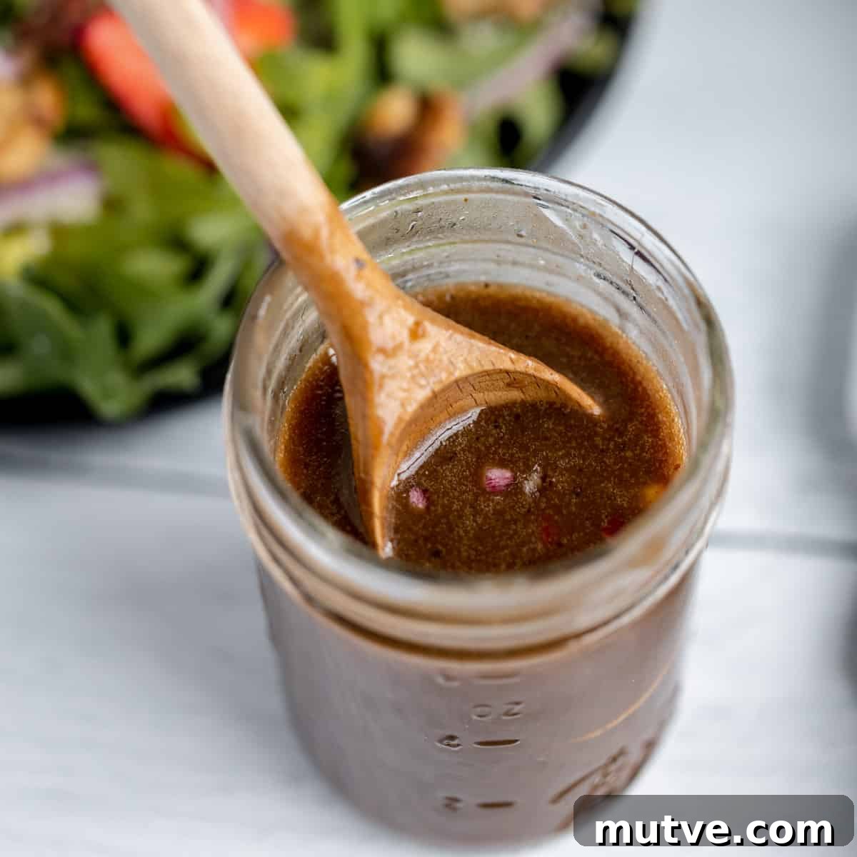 A jar of balsamic dressing with a wooden spoon. Salad in the background.