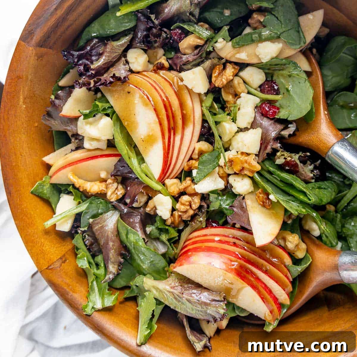A wooden bowl filled with a mixed green salad featuring apple slices, walnuts, cheese cubes, and a light dressing, with wooden salad servers.