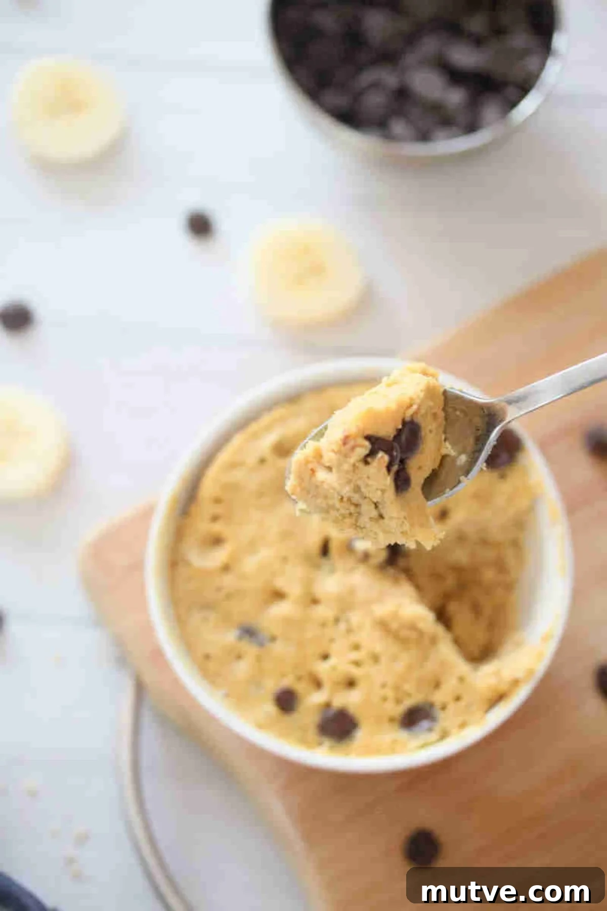 3-Ingredient Dairy-Free Protein Mug Cake 8 Mug cake with chocolate chips in a red bowl on a cutting board and a spoonful being lifted out.